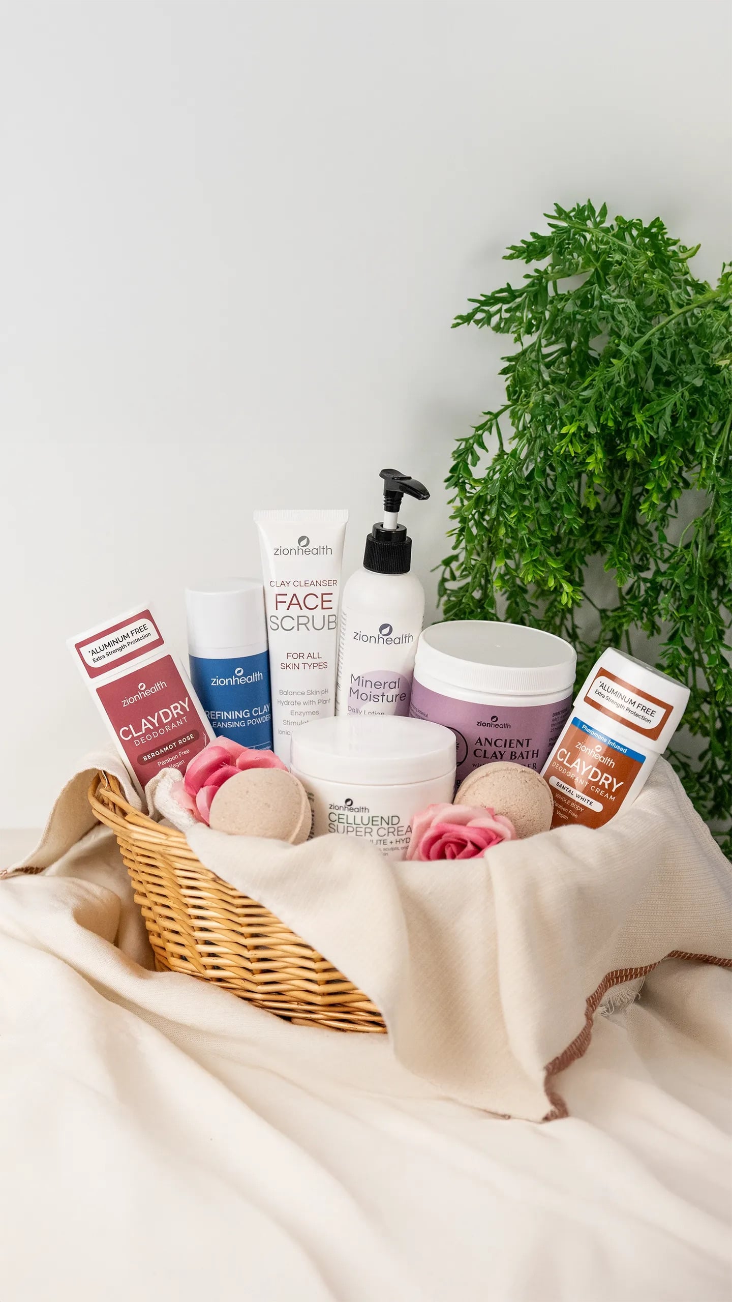 Zion Health skincare and body care products displayed in a wicker basket with clay deodorants, clay cleanser face scrub, mineral lotion, ancient clay bath, and cellulite cream, styled with pink roses and greenery on a soft neutral background.